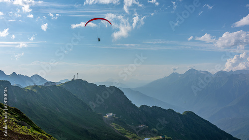 Red paraglider in blue cloudy sky over green mountains. Green valley with cable car down below. Krasnaya Polyana, Sochi, Caucasus, Russia.