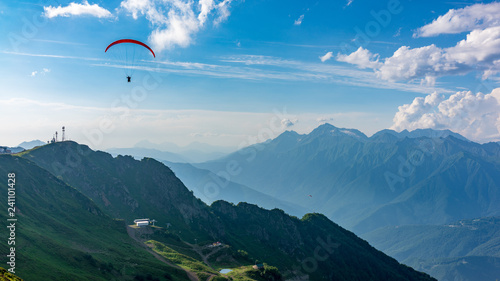 Red paraglider in blue cloudy sky over green mountains. Green valley with cable car down below. Krasnaya Polyana, Sochi, Caucasus, Russia.