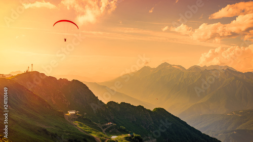 Red paraglider in the orange sunset cloudy sky over the green mountains. Green valley with cable car down below. Krasnaya Polyana, Sochi, Caucasus, Russia.