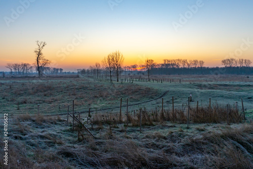 A winter scene with a path through a Dutch river delta landscape on the Belgian border. The rising sun colors the sky orange and blue.