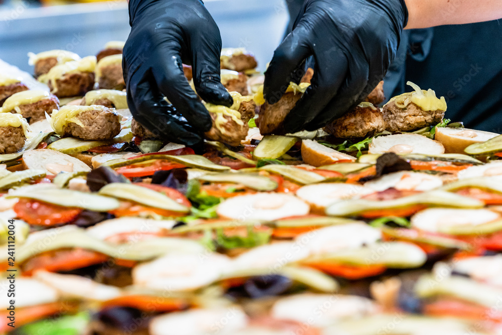 Female and Male Chef Putting Ingredients of Burgers on a Sliced Bread Spread on a Table in Black Gloves Female and Male Chef Putting Ingredients of Burgers on a Sliced Bread Spread on a Table in Black Gloves