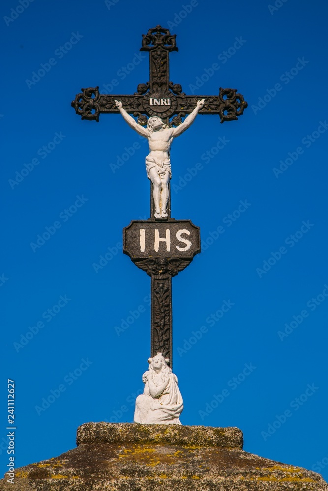 Christian black iron crucifix with white sculpture of Jesus Christ crucifixion, latin letters INRI, Iesus Nazarenus Rex Iudaeorum, Virgin Mary at the bottom of stone pedestal, sunny day, blue sky