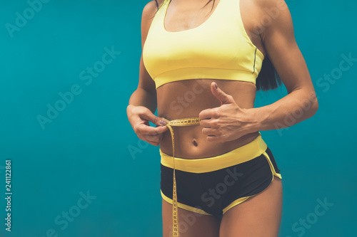 Crop shot of fitness woman in sport outfit measuring waistline with metric tape and showing thumb up. Good results of exercising in gym. Perfect shape of sportswoman. Achievments in body perfection. photo