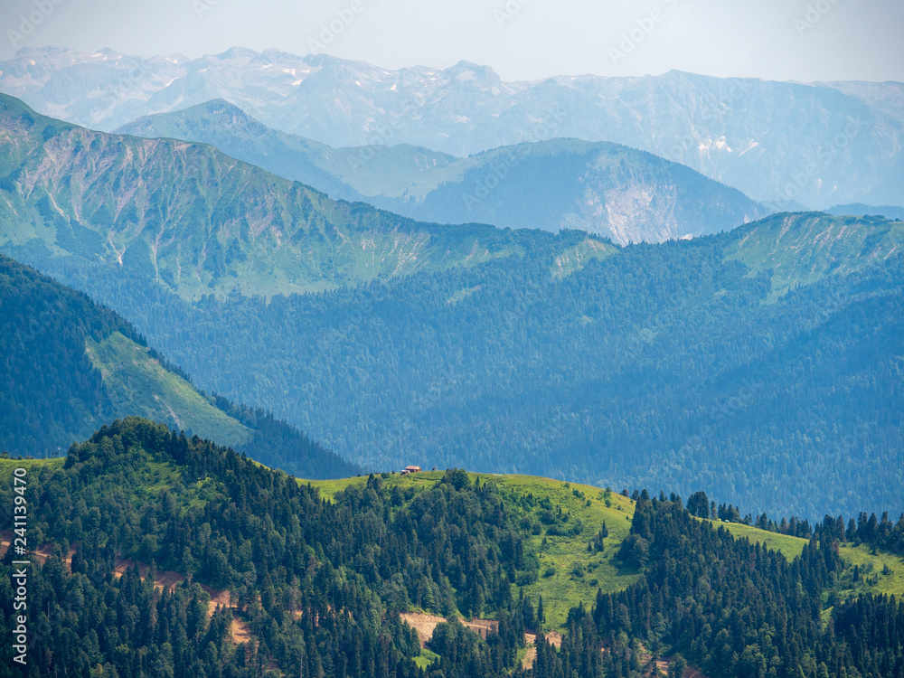 Small House on top of a green Ridge. Several high mountain ranges in the haze. Krasnaya Polyana, Sochi, Caucasus, Russia.