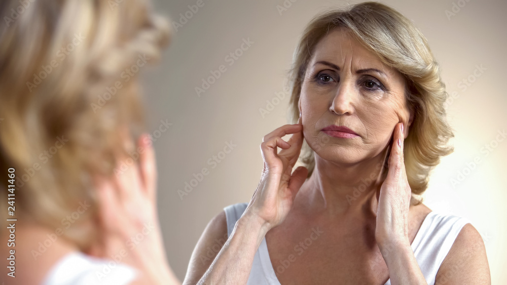 Unhappy aged woman looking in mirror at home, touching face, aging process Unhappy aged woman looking in mirror at home, touching face, aging process