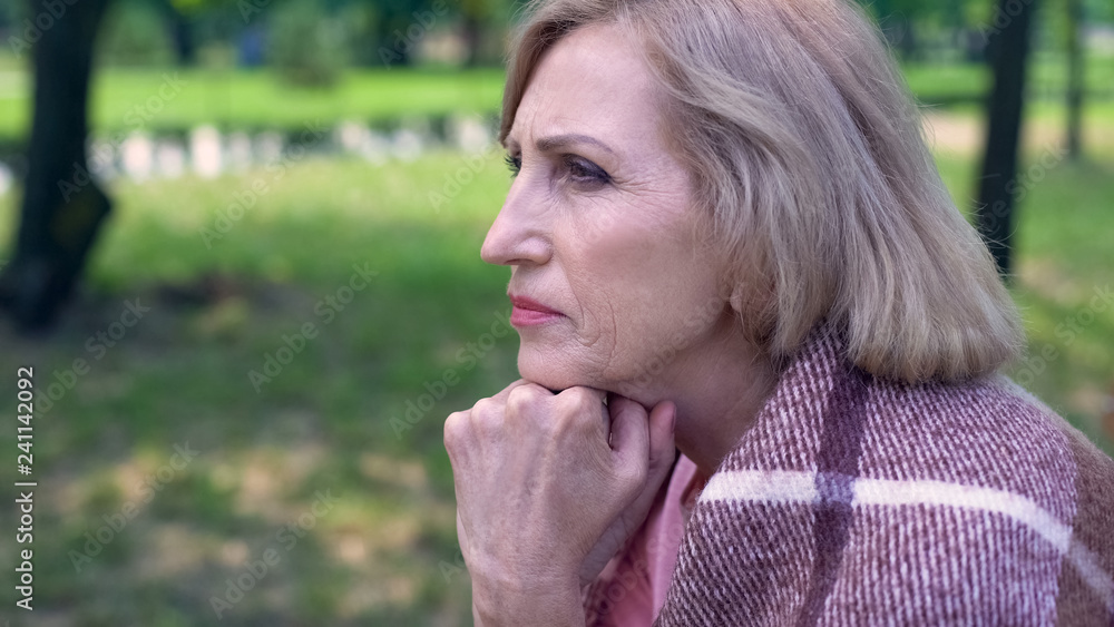 Thoughtful aged lady covered with plaid blanket sitting on bench in garden Thoughtful aged lady covered with plaid blanket sitting on bench in garden