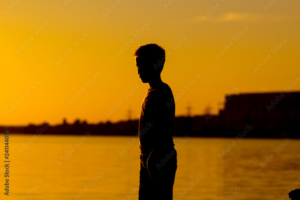 Fototapeta premium Silhouette of a boy standing near the river at sunset. Beach and calm water by the lake or river in the evening.