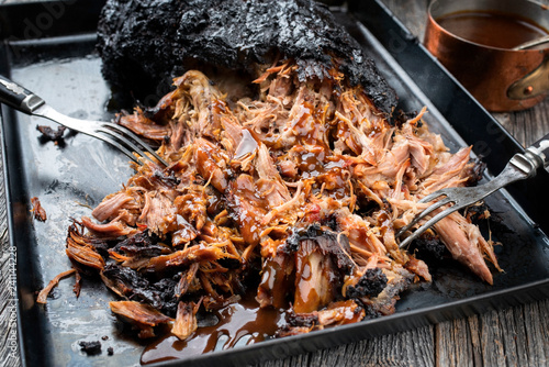 Traditional barbecue pulled pork piece of Bosten butt torn to bits with hot sauce in casserole as closeup on a board