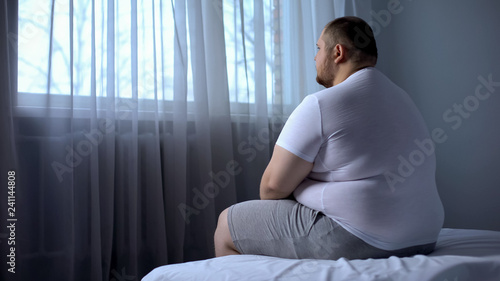 Sad heavy man sitting on bed at home, health problem, depression, insecurities photo