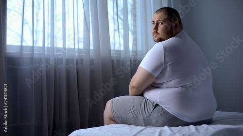 Sad fat man sitting on bed at home, looking at camera, depression, insecurities photo