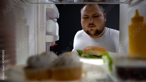 Bilde på lerret Overweight young man opening fridge at night to take big burger, calories