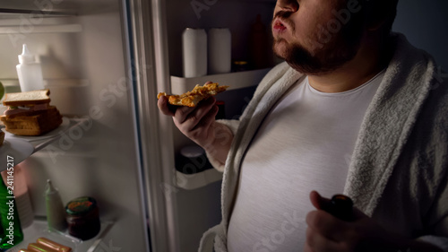 Obese bachelor eating pizza near fridge, holding beer, unhealthy lifestyle photo
