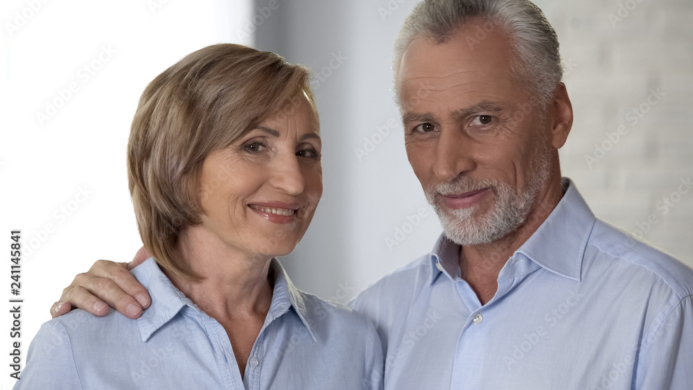 Smiling elderly couple looking at camera, man hugging lady, harmony in relations Smiling elderly couple looking at camera, man hugging lady, harmony in relations