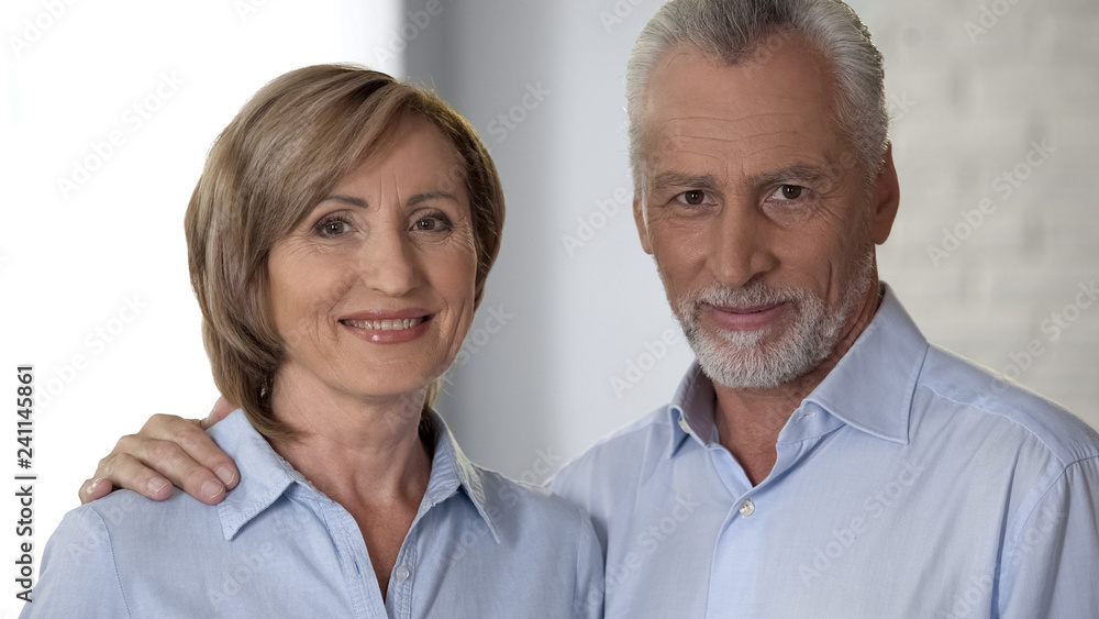 Happy elderly woman and man looking at camera, male hand lying on lady shoulder Happy elderly woman and man looking at camera, male hand lying on lady shoulder