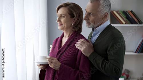 Картината върху платно Senior couple looking in window, male hugging lady shoulders from behind, family