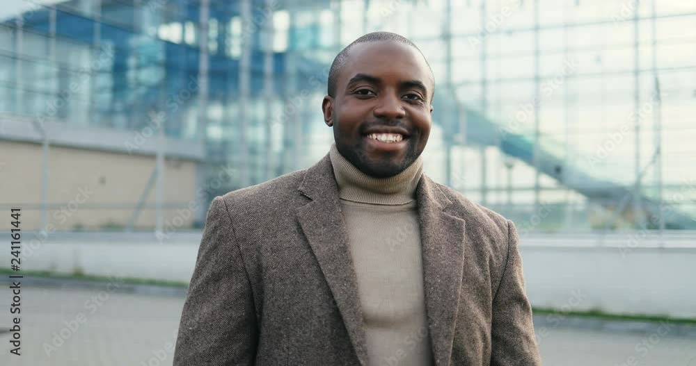 Portrait of the smiled young African American man in the casual style standing in front of the camera and looking at it on the big modern building background. Outdoor. Close up. Portrait of the smiled young African American man in the casual style standing in front of the camera and looking at it on the big modern building background. Outdoor. Close up.