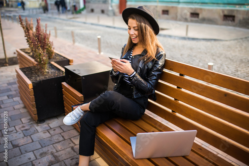 Pretty womanin fanky hat sitting on bench in city on summer day and reading text message on cell phone while using silver laptop and drinking takeaway coffee photo