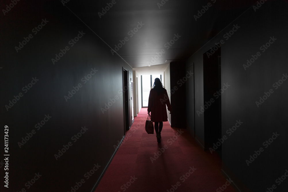 Girl walking through dark hallway in silhouette with bright light beaming through window.
