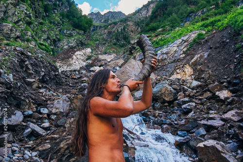 Fotografie Long haired bare-chested man blows horn in mountains