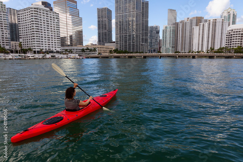 Adventurous girl kayaking in front of a modern Downtown Cityscape during a sunny evening. Taken in Miami, Florida, United States of America.