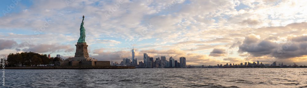 Panoramic view of the Statue of Liberty and Downtown Manhattan in the background during a vibrant cloudy sunrise. Taken in Jersey City, New Jersey, United States.
