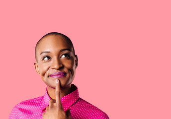 Portrait of a young woman in pink shirt with finger on mouth looking up thinking, isolated on pink studio background