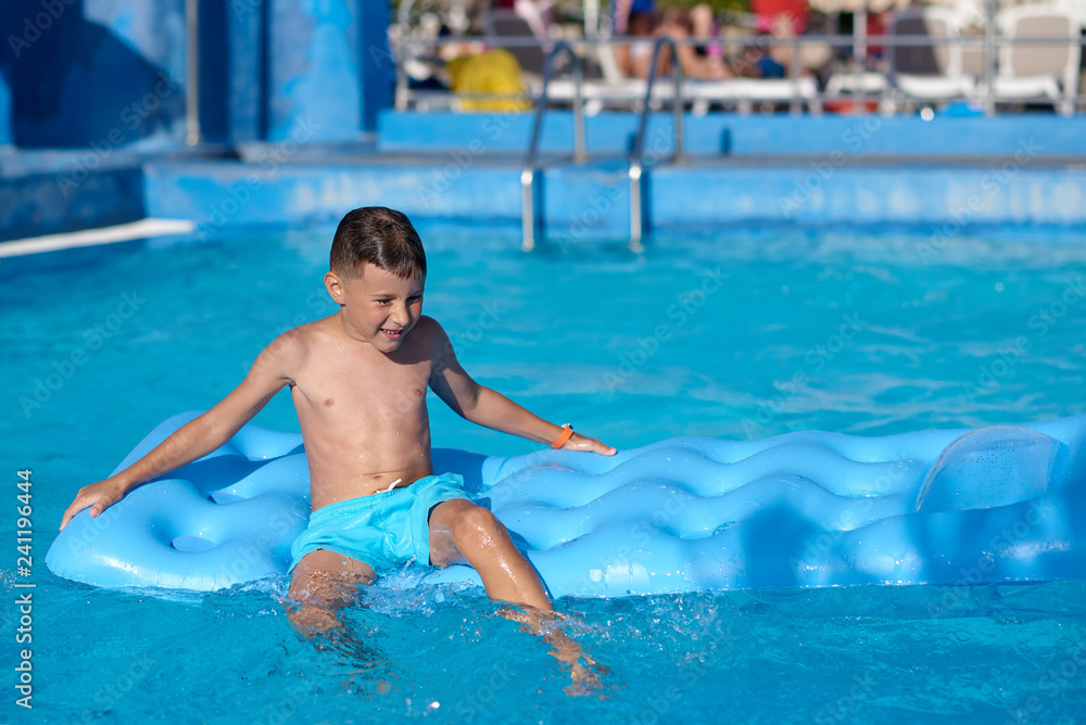 Caucasian boy is  sitting  on blue  inflatable mattress at hotel swimming pool. He is enjoying his summer vacations and smiling.