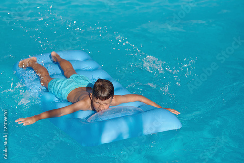  Caucasian boy is swimming on blue inflatable mattress at hotel swimming pool. He is enjoying his summer vacations. Copy space.
