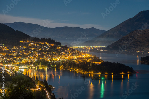 This photo was taken on top of a hill in Queenstown, New Zealand. It was night time and the town lights up beautifully. In the background, there are mountains. The lake reflects the lights.