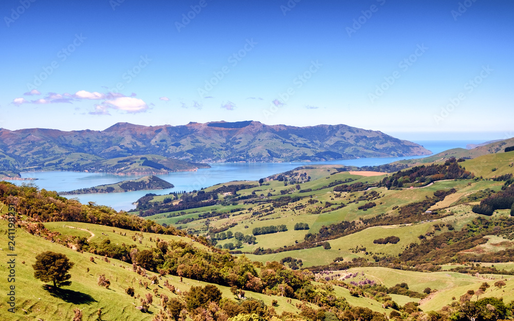 Beautiful travel image of Akaroa, New Zealand. This is popular retreat among tourist, travelers and locals. There are green hills, blue sea, farm, bay, peninsula, mountains and charming town.