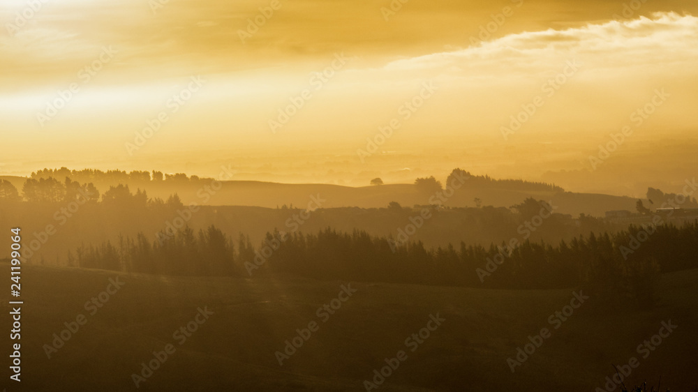 A beautiful sunrise image of trees and layers of mountains. The light is orange and warm. There are amazing light rays peaking through the trees. This image is suitable for background use.