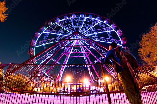 man stands in front of a glowing illumination by a ferris wheel  reflected in a puddle