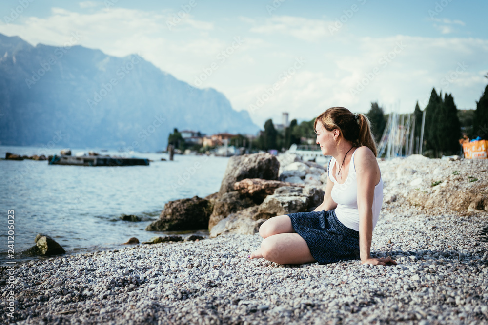 Beautiful young woman is sitting on the pebble beach and enjoying the view, vacation