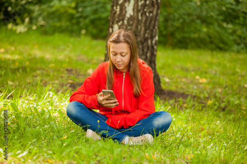 modern teenager girl sitting on the grass in the park with a smartphone in her hands © izida1991