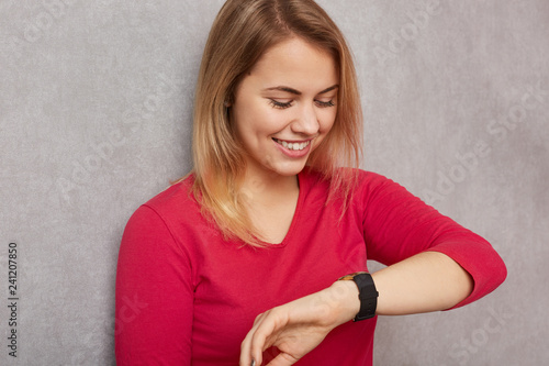 Image of cheerful blonde woman looks positively at wristwatch, checks time, rejoices having spare time before meeting, dressed in red jumper, isolated over grey background. People and time concept