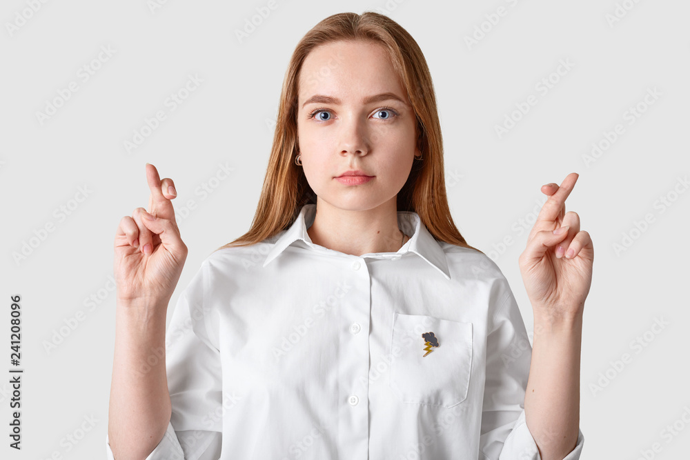 Young pretty female crosses fingers, has blue eyes, dressed in formal white shirt, has long hair, wishes good luck, isolated over white background. Serious student prays for dreams come true Young pretty female crosses fingers, has blue eyes, dressed in formal white shirt, has long hair, wishes good luck, isolated over white background. Serious student prays for dreams come true