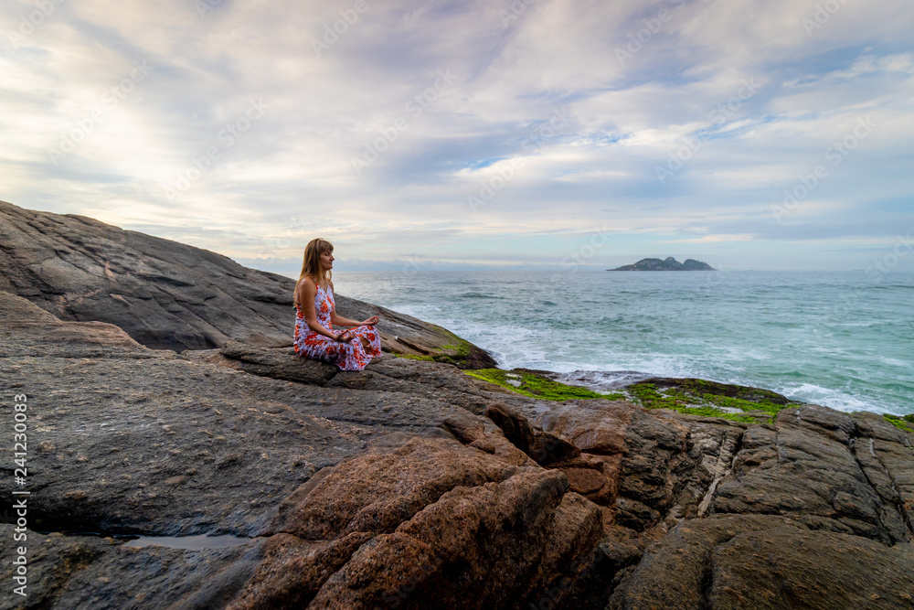 Landscape with a woman in a meditation pose on a rock at the waterfront of an ocean coast during a beautiful coloured sunrise with an island in the background