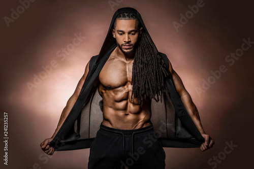 Muscular African American Black athletic fitness model wearing  black hoodie with six pack abs in studio with dramatic lighting against a brown background  