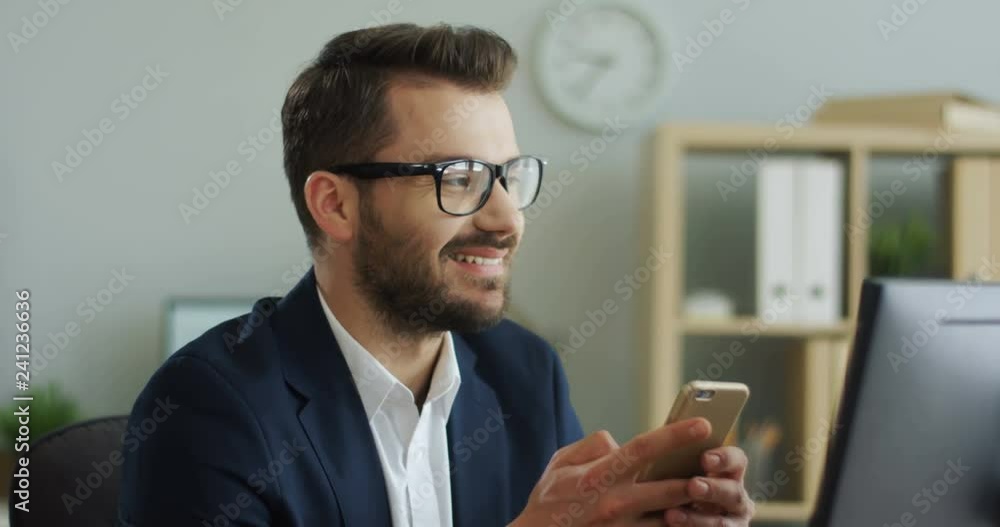 Portrait of the young attractive smiled Caucasian businessman in glasses using smartphone and chatting on it in the office. Close up. Portrait of the young attractive smiled Caucasian businessman in glasses using smartphone and chatting on it in the office. Close up.