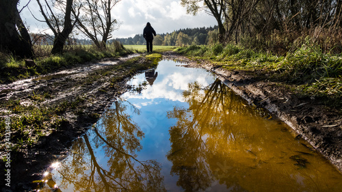 Human figure on off-road track. Tree mirroring in water close-up. Man silhouette. Muddy bumpy field path in spring landscape. Forest in background. Reflection of a blue sky and white clouds in puddle. © KPixMining