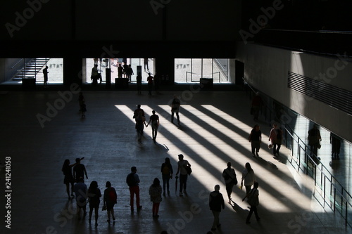 Interior of urban art gallery vast open space with people moving walking through, columns of light streaming white as blocks against dark shadow with figures silhouetted in Tate Modern entrance London photo
