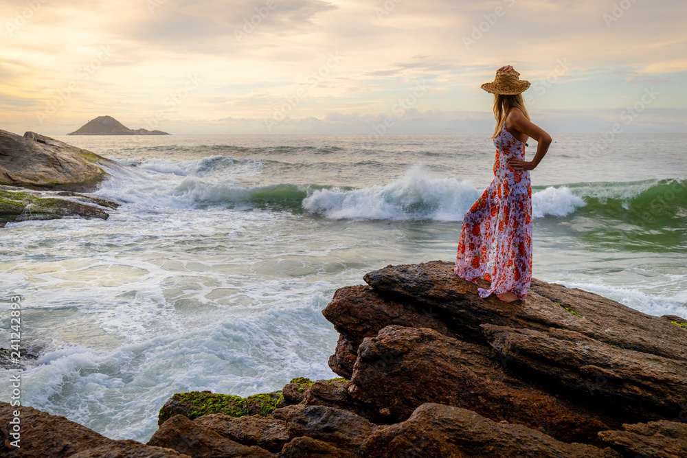 Beautiful slender woman with a hat and stylish dress standing on a beach with the waves coming in at sunrise Beautiful slender woman with a hat and stylish dress standing on a beach with the waves coming in at sunrise