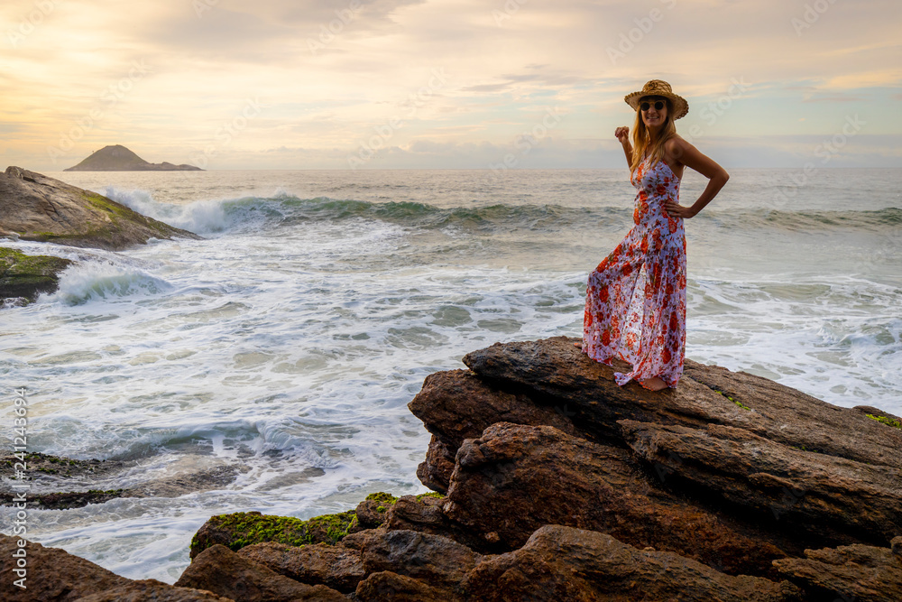 Beautiful slender woman in stylish dress and sunglasses standing standing on a rock with the waves rolling in from the wider seascape at sunrise Beautiful slender woman in stylish dress and sunglasses standing standing on a rock with the waves rolling in from the wider seascape at sunrise