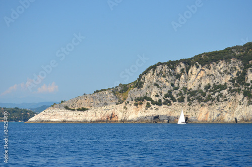 A yacht with a white sail is on the blue waters of the Mediterranean Ocean. Behind is a rocky island, dotted with scrub. The mainland with hills is in the distance. The sky is blue.