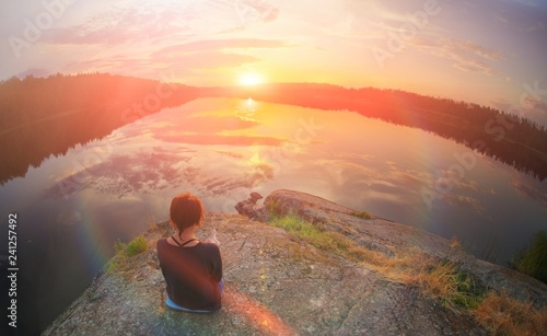 Young woman sitting enjoying peaceful moment of beautiful colorful sunset. In the reflection of the lake water sees clouds and sun. Vintage mood, concepts of winner, freedom, happiness.