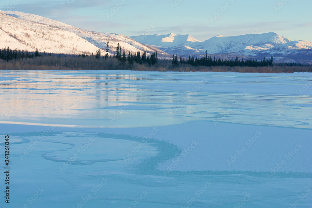 Snowy mountains in the rays of dawn. Winter landscape with the mountains and the frozen lake in Yakutia, Siberia at sunrise. Clear blue ice of the frozen lake at morning Frosty morning, day or evening