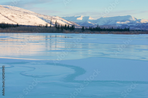 Snowy mountains in the rays of dawn. Winter landscape with the mountains and the frozen lake in Yakutia, Siberia at sunrise. Clear blue ice of the frozen lake at morning Frosty morning, day or evening