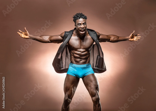 Muscular African American Black athletic man screams at the camera with hands open wearing blue underwear with six pack  abs in studio with dramatic lighting against a brown background  