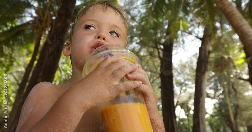 Adorable little kid drinking cold beverage fruit fresh and looking away while standing on blurred background of tropics Adorable little kid drinking cold beverage fruit fresh and looking away while standing on blurred background of tropics