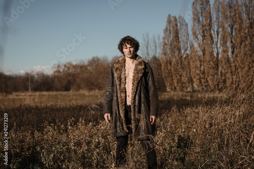 un ragazzo a torso nudo indossa una pelliccia in piedi in un prato selvaggio. Lui ti guarda © deversteel